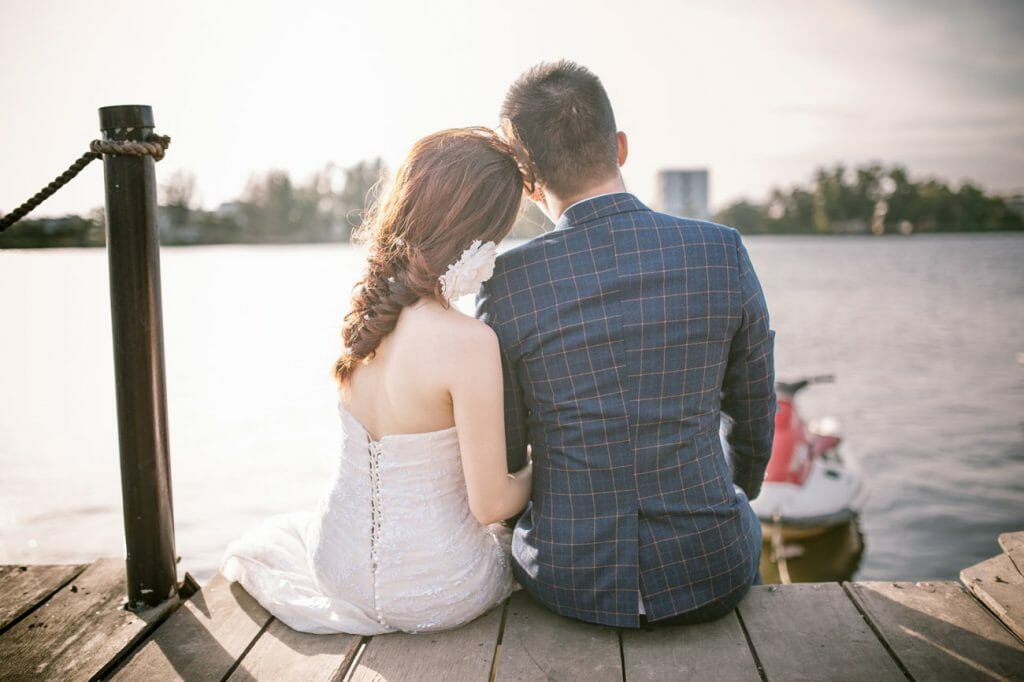 A shot of the back of a bride and groom, sat on a pier, overlooking a river