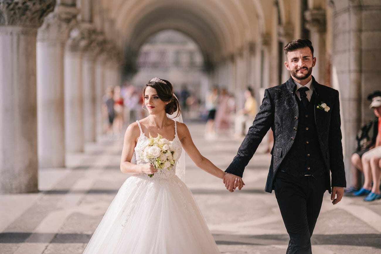 A bride and groom holding hands under stone arches, walk towards the camera