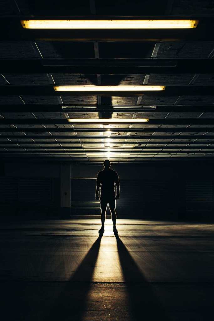 A man standing underneath a line of lights which creates a line of shadows as well.