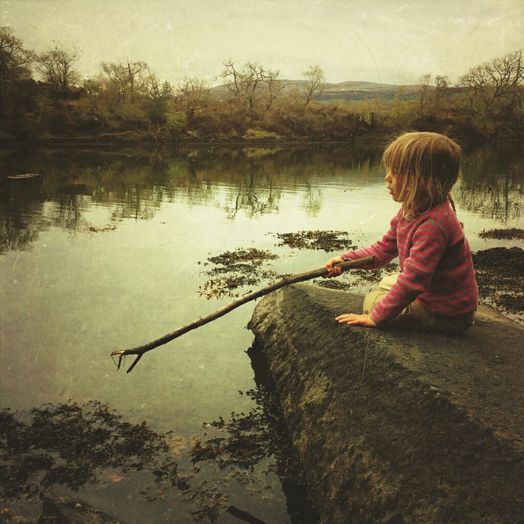 A mobile photography photo of a child sat on a rock, about to dip a stick in a lake