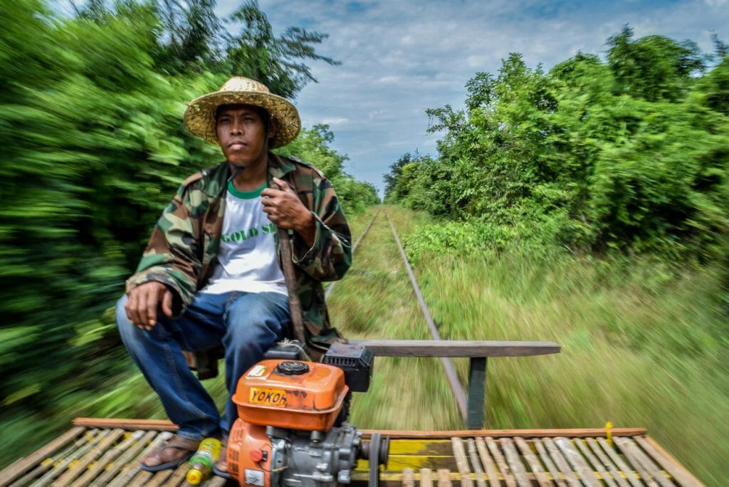 Man sits on top of a bamboo train while he hurtles down tracks as the scenery around him shoots past as a green motion blur