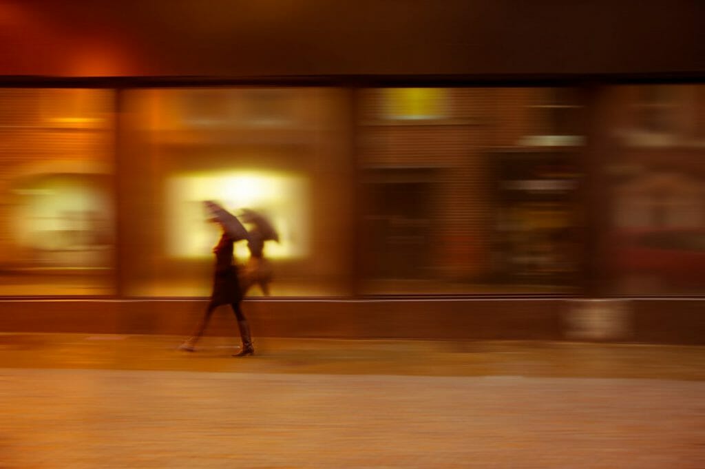 A hazy figure under an umbrella makes her way down a street which has been blurred with motion blur