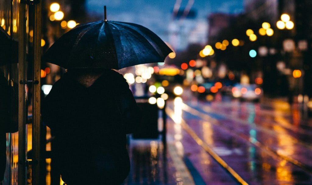 A full frame photo night shot of a person under an umbrella looking out at a wet street