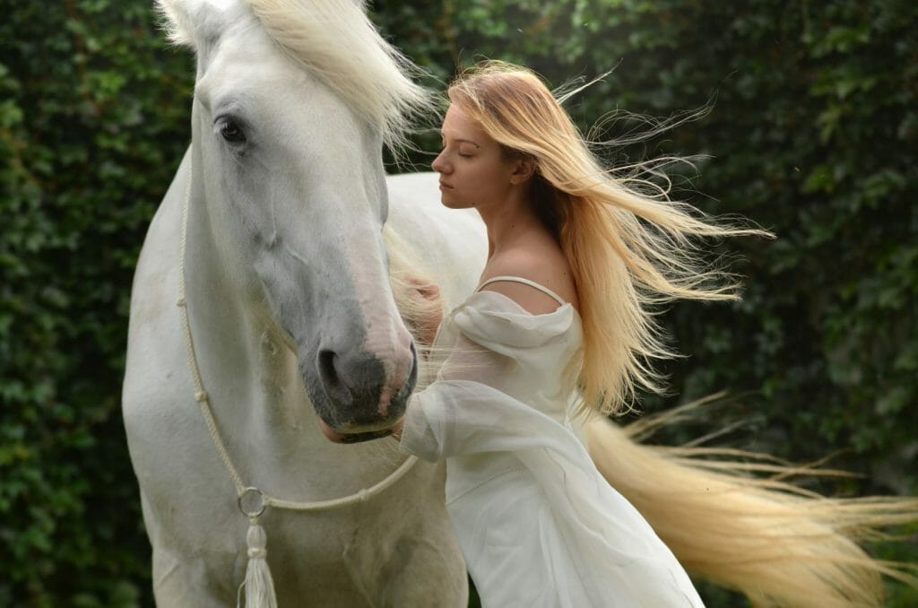 A photo of a woman wearing a white dress stood next to a white horse