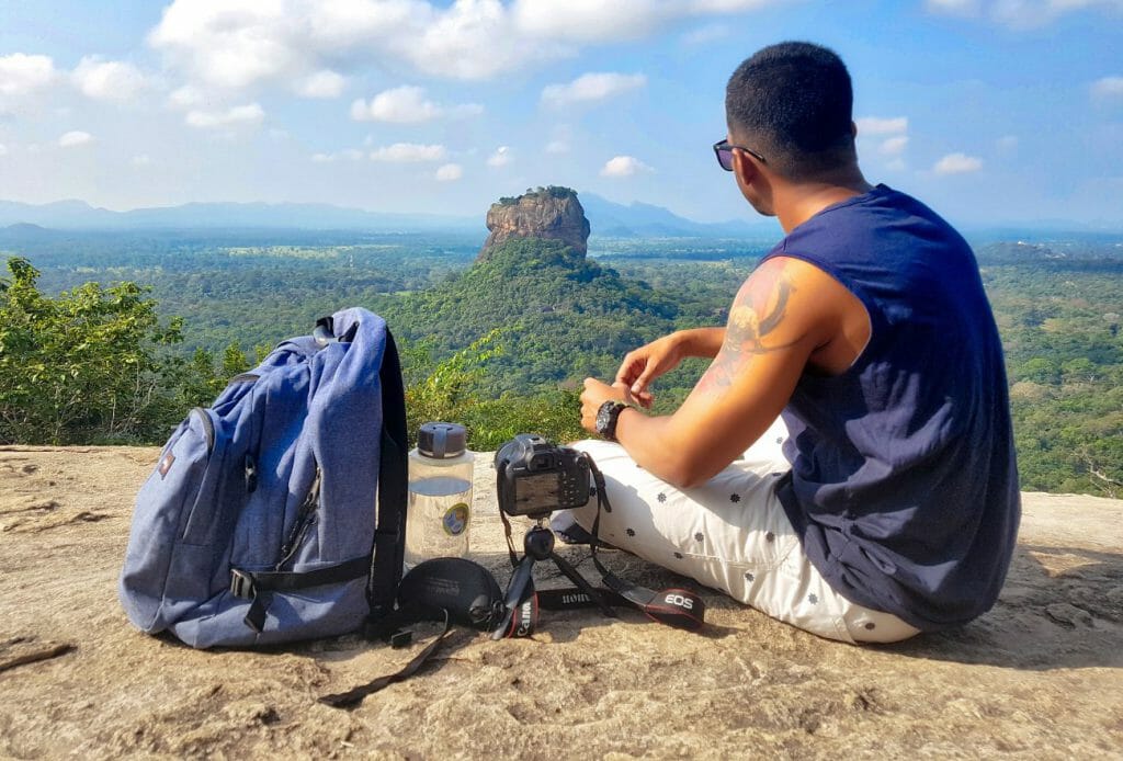 A man sits with a camera and tripod looking out over hills and cliffs