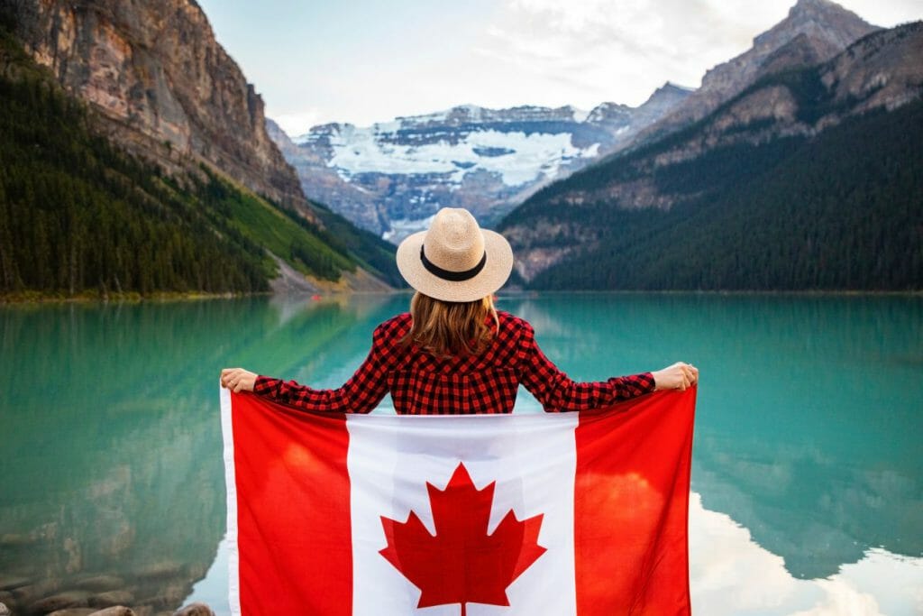 A woman with a Canadian flag stood on the side of a lake looking at mountains