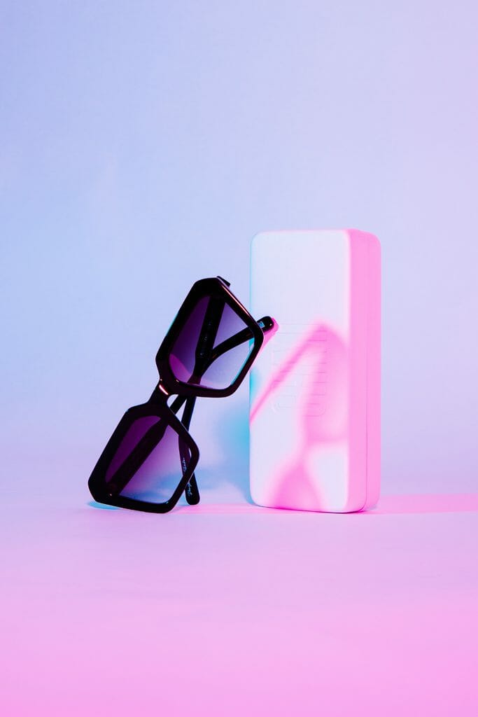 A pair of black sunglasses leaning against a white case on a pink/purple background