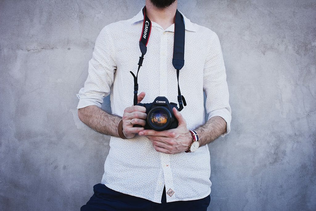 A man stands against a wall holding a DSLR camera