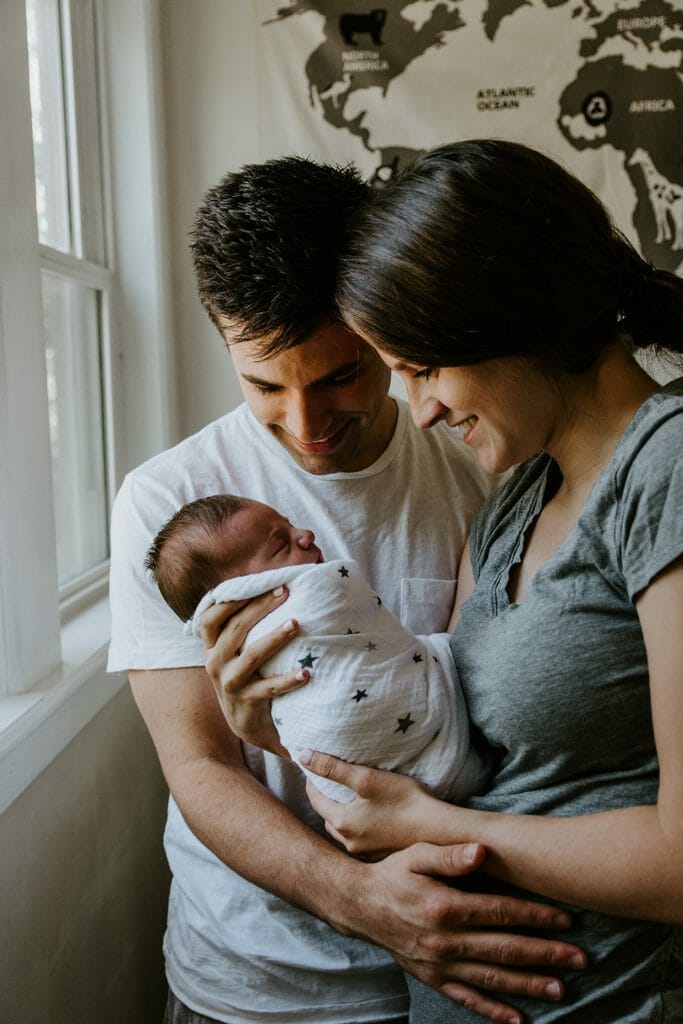 An indoor photography image of parents gazing down at their newborn baby, lit by natural light from a nearby window