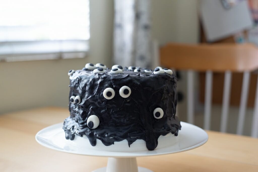 A black cake with eyes on it sat on a cake stand on a table