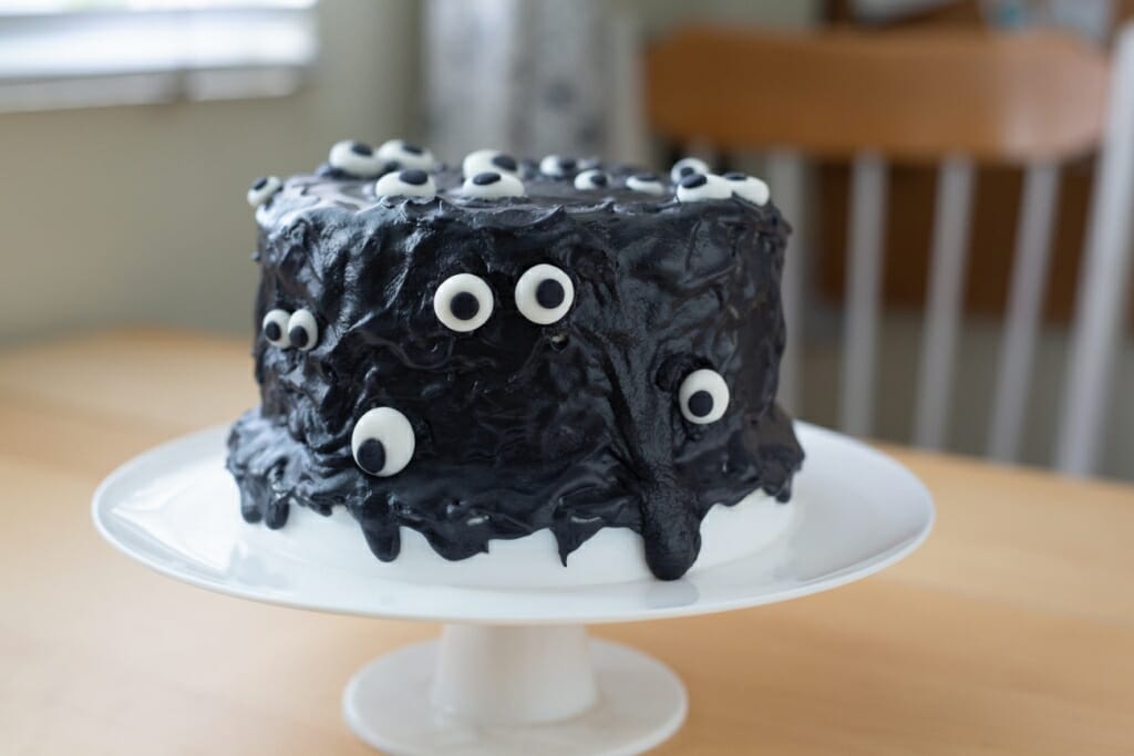 A black cake with eyes sat on a cake stand on a table