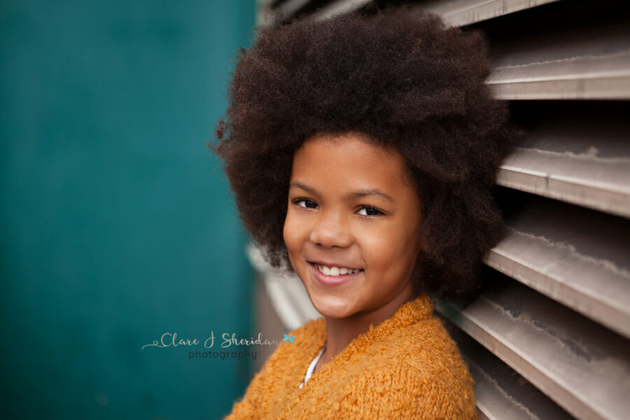 A smiling girl wearing an orange cardigan in front of a turquoise wall - a great example of kids' photography