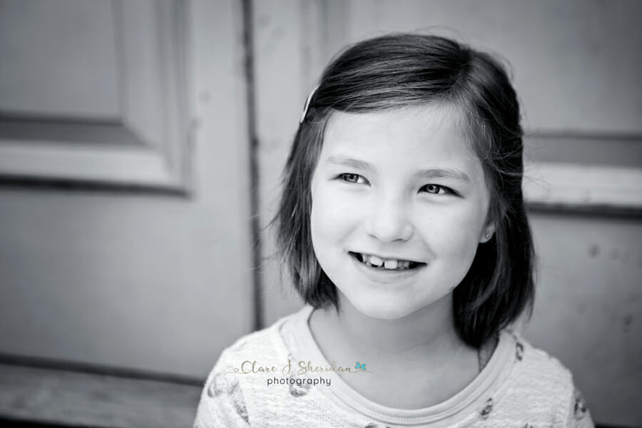 A black and white shot of a smiling girl sat in front of a door - another example of kids' photography