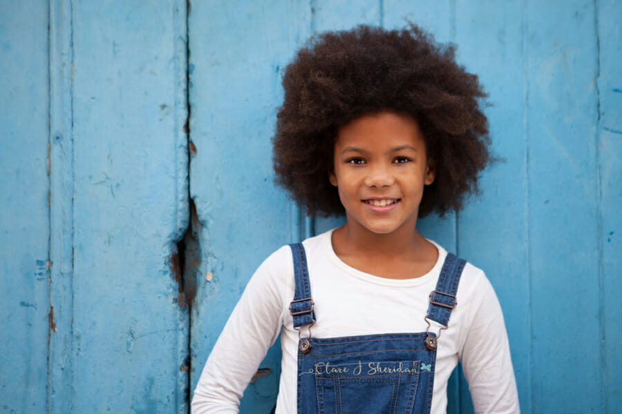 A girl in dungarees stood in front of blue wood - another example of kids' photography