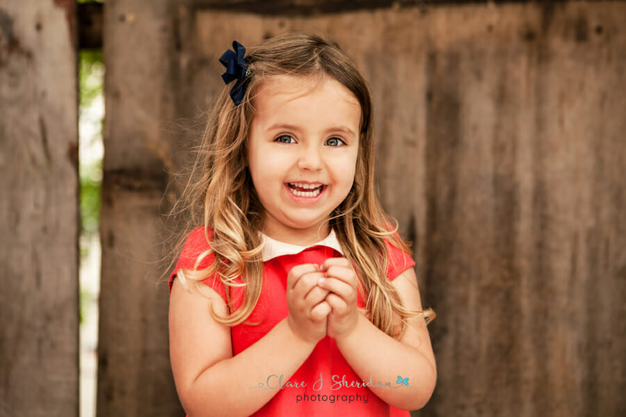 A laughing girl in a red dress stood in front of a wooden fence - another example of kids' photography