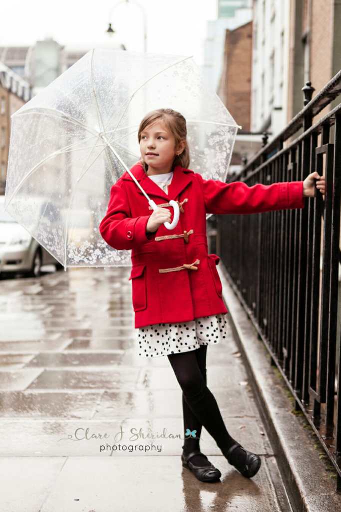 A girl in a red duffel coat holds onto railings under an umbrella on a rainy day