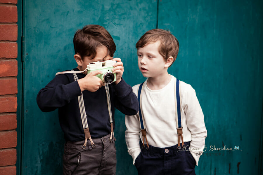 Two young boys play with a camera in front of a turquoise wall