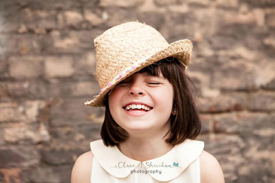 A girl wearing a summer hat laughing in front of a brick wall