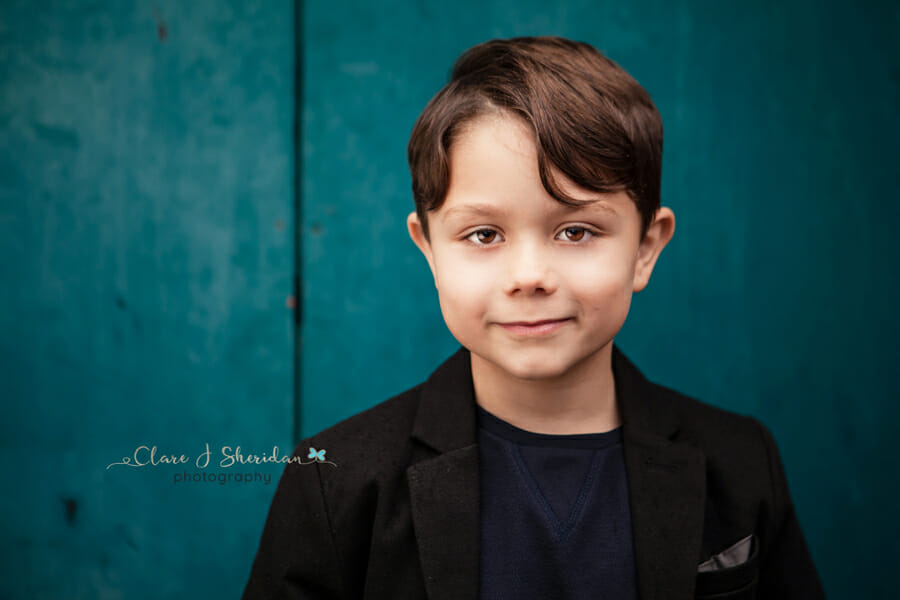 A young boy in a black blazer stood in front of a turquoise background