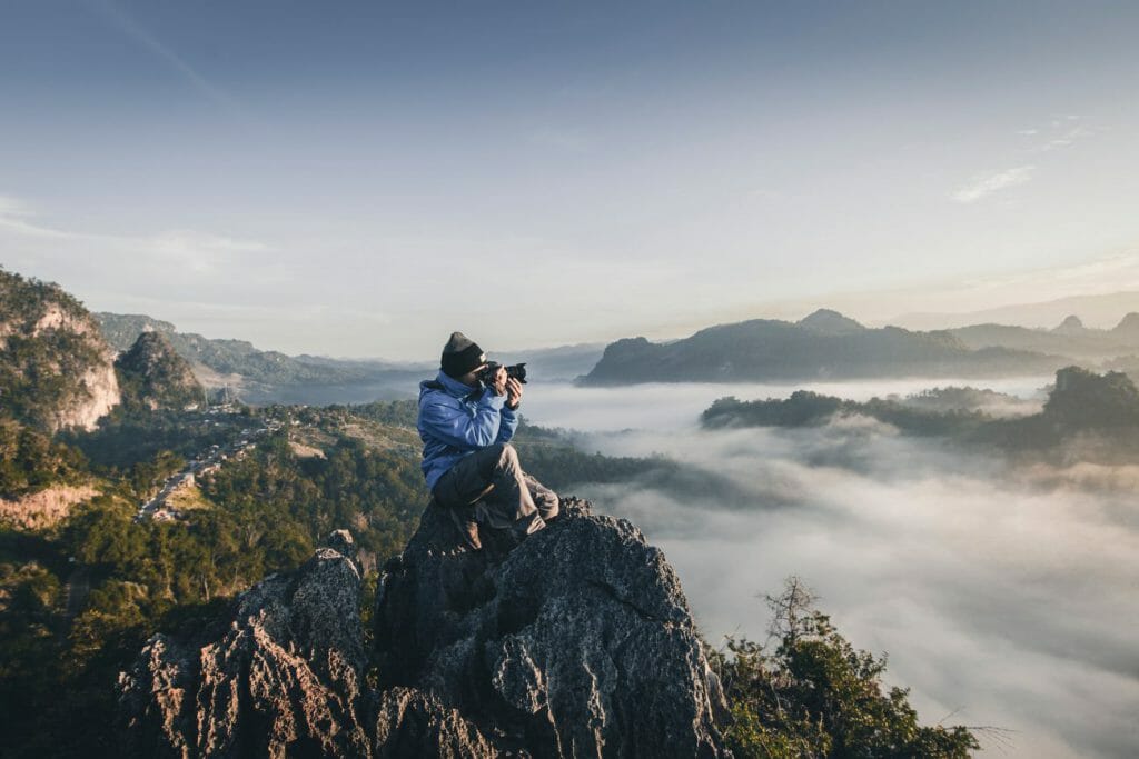 A travel photography shot of a man sat on a cliff, taking photos of misty mountains