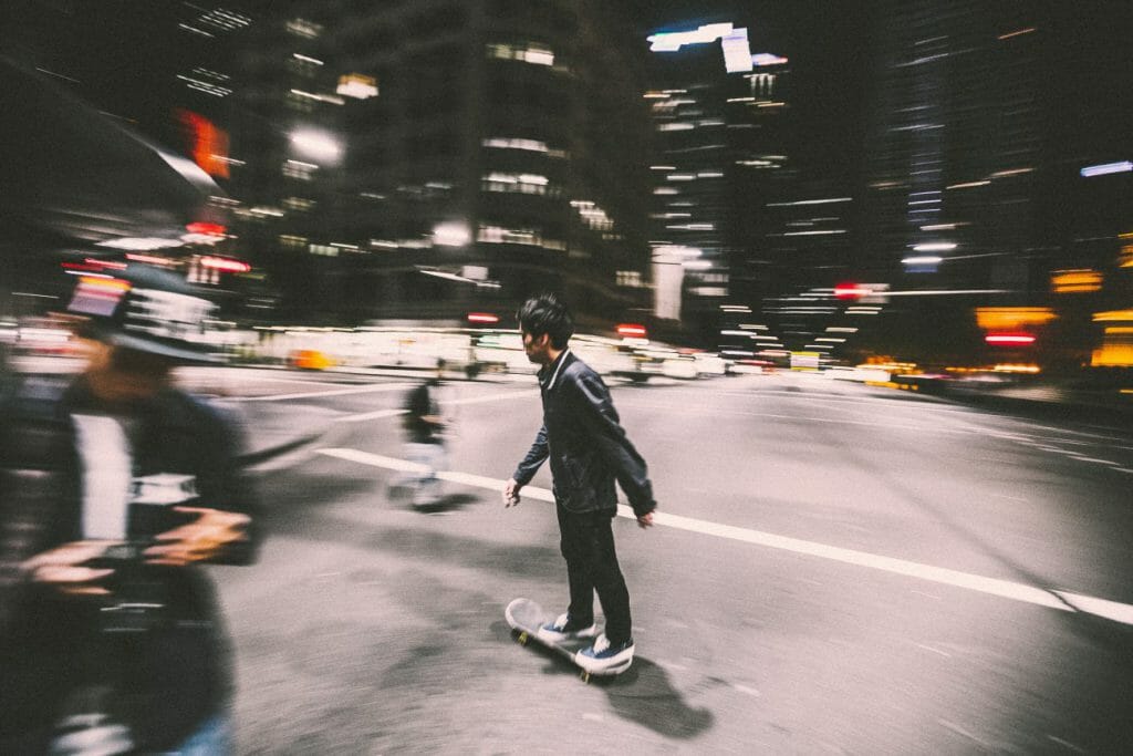 A panning shot of a man skateboarding through a city at night